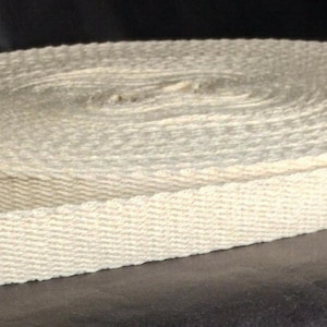 May include: A roll of off-white, woven cotton webbing. The webbing is tightly wound in a circular shape, with a portion unrolled, revealing its flat, rectangular form. The material has a textured surface and a natural, neutral color.