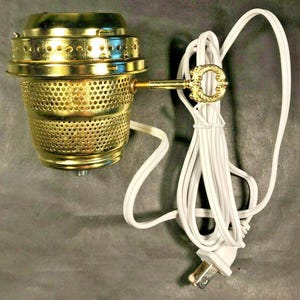 May include: A gold-colored, perforated metal lamp with a key-shaped accent. The lamp has a white electrical cord and plug. The lamp's design includes a perforated shade and a decorative key detail.