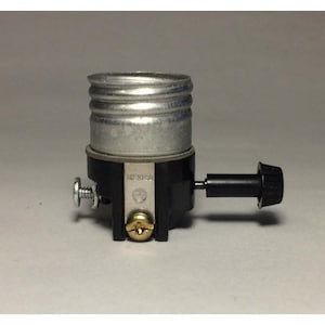 Könnte beinhalten: Eine silberfarbene Lampenfassung mit schwarzem Sockel und schwarzem Knopf. Die Fassung hat eine Schraube und eine Metallplatte mit dem Wort "MEXICO". Die Lampenfassung ist für den elektrischen Gebrauch bestimmt.