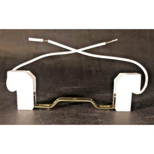 May include: A white electrical component with two white ceramic bulb holders, a gold-colored metal bracket, and white wires. The wires cross over each other. The background is dark.