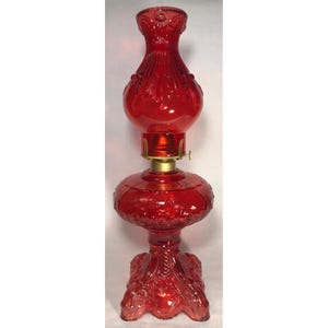 May include: A vibrant red glass oil lamp featuring an ornate embossed pattern across the base and the chimney. The lamp includes a brass-colored metal burner assembly connecting the bulbous reservoir to the flared top chimney section.