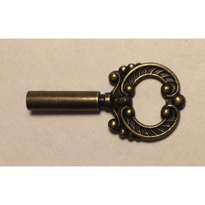 May include: An antique-style key with a bronze finish. The key features an ornate, circular head with decorative detailing, including small spheres and leaf-like patterns. The shaft is cylindrical and solid.