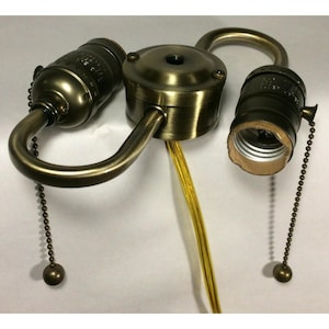 May include: A vintage-style brass lamp fixture with two bulb sockets. The fixture has a central mounting plate, curved arms, and pull chains with decorative ball ends. A yellow electrical cord extends from the center.