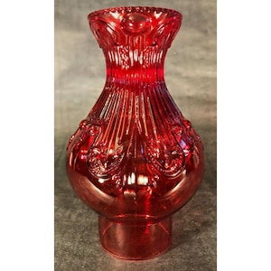 May include: A ruby red glass vase with a bulbous body and a flared, ornate top. The vase features vertical ribbing and detailed scrollwork. The base is a short, cylindrical shape. The glass has a glossy finish, reflecting light.