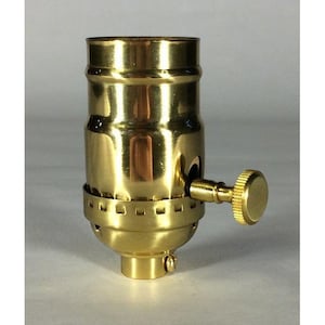 Könnte beinhalten: Eine Lampenfassung aus poliertem Messing mit zylindrischem Körper und glatter, reflektierender Oberfläche. Das Bauteil verfügt über einen seitlich angebrachten, gerändelten Drehknopf zur Bedienung sowie einen Gewindesockel mit kleinen Lüftungsschlitzen.
