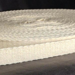 May include: A roll of off-white, woven cotton webbing. The webbing is tightly wound in a circular shape, with a portion unrolled and extending outward. The texture of the material is visible, showing the weave pattern. The background is dark.