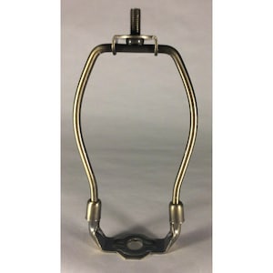 May include: A brass-toned metal lamp harp with a curved frame and a threaded finial stud at the top. The base features a circular opening for attachment, and the structure is designed to support a lampshade on a standard table or floor lamp.