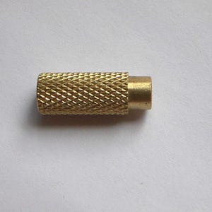 May include: A small, cylindrical brass component with a diamond-patterned grip. The component has a smooth, smaller cylinder extending from one end. The item is likely a hardware part or a small tool component.