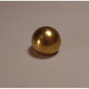 Puede incluir: Una pequeña esfera dorada y sólida. La bola tiene una superficie lisa y reflectante, que capta la luz y crea reflejos. El objeto se encuentra sobre un fondo neutro, enfatizando su forma y color.