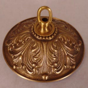 May include: Ornate brass ceiling canopy with a detailed, swirling leaf design. The canopy has a central loop for hanging and a decorative beaded edge. The overall finish is a warm, antique gold color, suitable for vintage or classic decor.