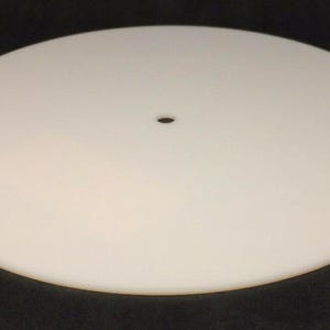 May include: A white, circular object with a small hole in the center. The object is smooth and opaque, and appears to be made of a solid material. The background is a dark color, providing contrast to the white disc.