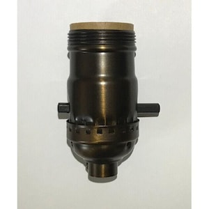 May include: A bronze-colored lamp socket with a brown cardboard insert at the top. The socket has a cylindrical body with a series of horizontal ridges and two small, rectangular side pieces. The base has a smaller diameter and a rounded bottom.
