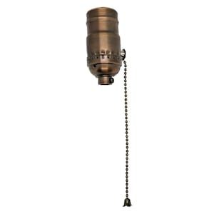 Peut inclure: Une douille de lampe cylindrique avec une finition en bronze brossé, présentant une texture nervurée verticale sur la partie inférieure et une longue chaînette à billes en métal assortie, se terminant par un petit embout décoratif en forme de fleur.