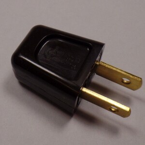 May include: A black electrical plug with two brass-colored prongs. The plug has a rectangular shape with rounded edges and a small indentation on the top. The prongs have small holes near the tips.