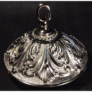 May include: A polished silver-tone metal canopy for a light fixture, featuring a domed shape with an intricate embossed acanthus leaf pattern and a central loop at the top for suspension, showcasing a reflective finish and textured detailing.
