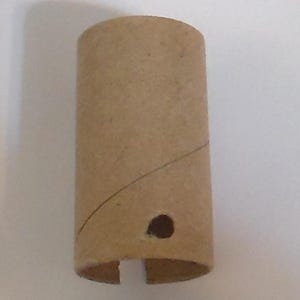 May include: A cylindrical cardboard tube, tan in color, with a small circular hole and a curved line. The bottom edge has two cut-out sections. The tube is standing upright against a white background.