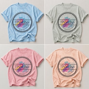 Puede incluir: Cuatro camisetas de manga corta en diferentes colores: azul claro, gris, rosa y melocotón. Cada camiseta tiene un diseño circular con el texto "TINYLINY" y "MOCKS" en un marco negro. Las camisetas tienen un aspecto texturizado y ligeramente descolorido.