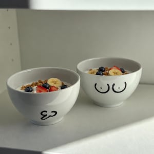 Può includere: Due ciotole in ceramica bianca riempite con yogurt, granola, fette di banana, fragole e mirtilli. La ciotola anteriore presenta un disegno a linee nere a forma di cuore, mentre quella posteriore mostra un disegno a linee nere di due cerchi.