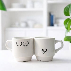 Può includere: Due tazze in ceramica bianca con illustrazioni a tratto nero. Una tazza presenta un paio di seni, l'altra un pene. Le tazze sono ambientate su uno sfondo bianco sfocato, che suggerisce una cucina o una sala da pranzo.