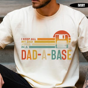 May include: Ivory-colored t-shirt with the text "I KEEP ALL MY DAD JOKES IN A DAD-A-BASE" in a retro design. The graphic includes a floppy disk and a sunset-like color scheme of orange, yellow, and teal. The shirt is a casual wear item.