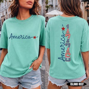 May include: A mint green short-sleeve t-shirt featuring the word America in dark blue script with a red star on the front. The back displays the word America vertically in blue script next to the number 1776 in red, white, and blue flag patterns.