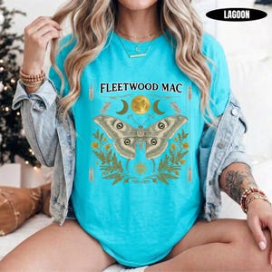 Może przedstawiać: Jasnoturkusowy t-shirt z nadrukiem graficznym i napisem FLEETWOOD MAC. Wz&oacute;r przedstawia dużą ćmę nad ż&oacute;łtymi liśćmi paproci, otoczoną p&oacute;łksiężycami i pełnią księżyca, z małym symbolem oka umieszczonym w dolnej części grafiki.