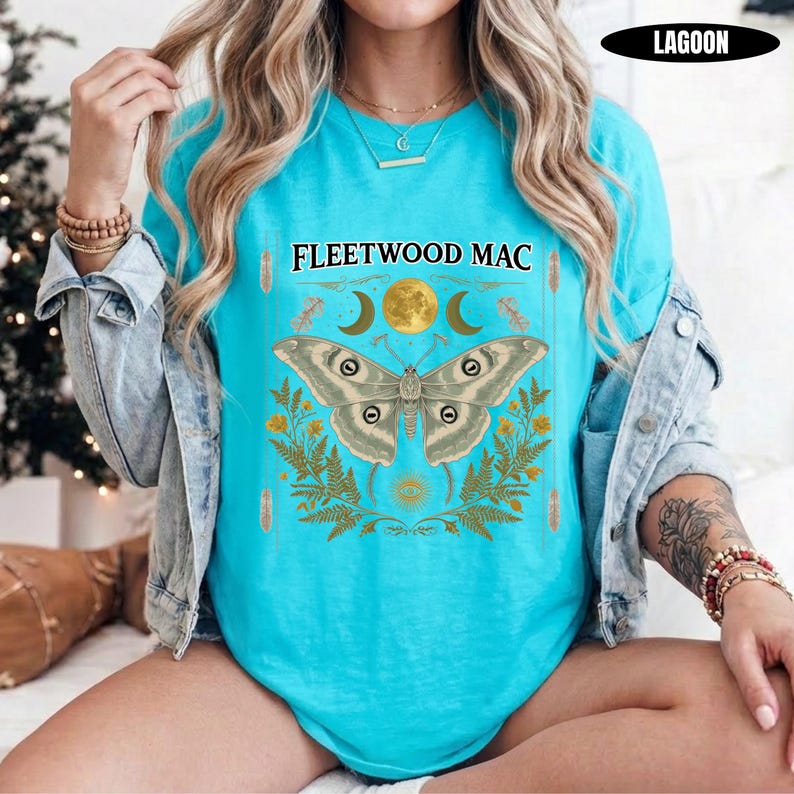 Może przedstawiać: Jasnoturkusowy t-shirt z nadrukiem graficznym i napisem FLEETWOOD MAC. Wz&oacute;r przedstawia dużą ćmę nad ż&oacute;łtymi liśćmi paproci, otoczoną p&oacute;łksiężycami i pełnią księżyca, z małym symbolem oka umieszczonym w dolnej części grafiki.