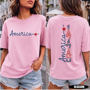 May include: A light pink short-sleeve t-shirt featuring the word America in dark blue script with a red star on the front. The back displays the word America and the number 1776 in a vertical design with red, white, and blue flag patterns.