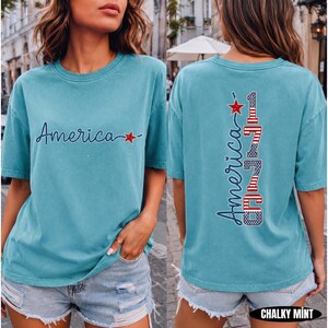May include: A chalky mint short-sleeve t-shirt featuring the word America in dark blue script with a red star on the front. The back displays the word America and the number 1776 in a vertical design with red, white, and blue flag patterns.