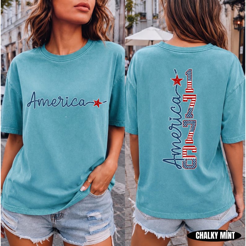 May include: A chalky mint short-sleeve t-shirt featuring the word America in dark blue script with a red star on the front. The back displays the word America and the number 1776 in a vertical design with red, white, and blue flag patterns.