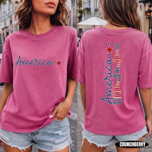 May include: A dusty rose short-sleeve t-shirt featuring the word America in blue script with a red star on the front. The back displays the word America in blue script above the vertical text 1776, styled with a red, white, and blue flag pattern.