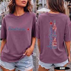 May include: A berry-colored short-sleeve t-shirt featuring the word America in blue script with a red star on the front. The back displays the word America vertically alongside the number 1776, styled with red, white, and blue flag patterns.