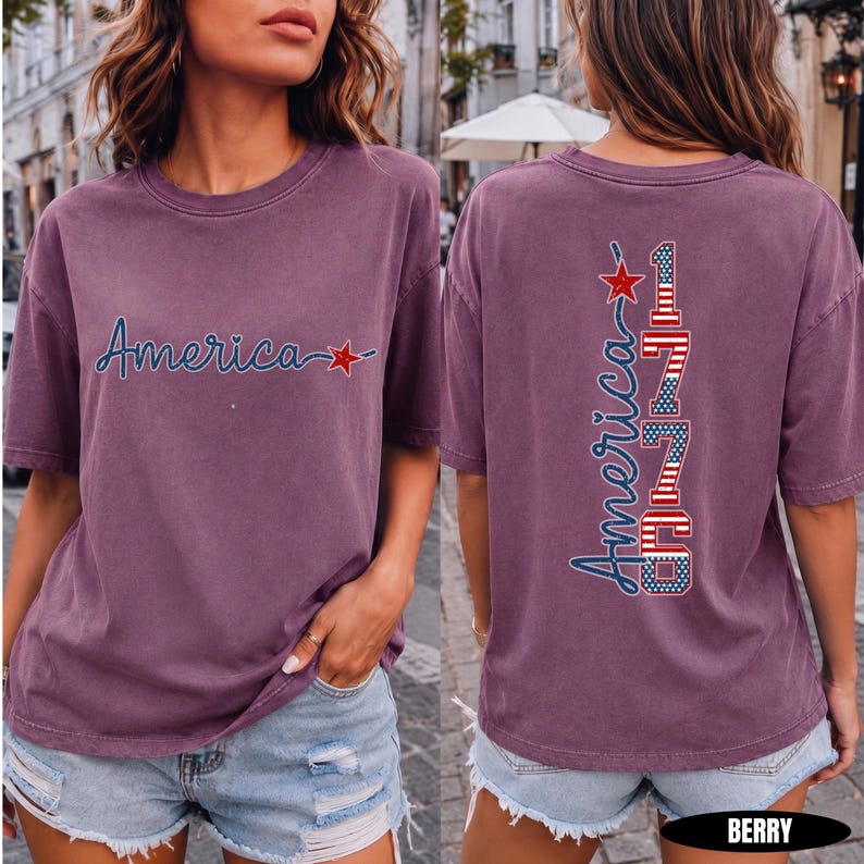 May include: A berry-colored short-sleeve t-shirt featuring the word America in blue script with a red star on the front. The back displays the word America vertically alongside the number 1776, styled with red, white, and blue flag patterns.