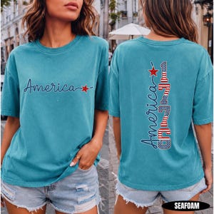 May include: A seafoam green short-sleeve t-shirt featuring the word America in cursive script with a red star on the front. The back displays the word America vertically alongside the number 1776, styled with red, white, and blue flag patterns.