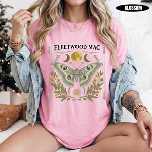 Może przedstawiać: Jasnor&oacute;żowy t-shirt z napisem Fleetwood Mac nad grafiką przedstawiającą dużą ćmę, pełnię księżyca i liście paproci. Wz&oacute;r zawiera symbole niebieskie i ozdobne linie. Koszulka jest noszona pod jasnoniebieską kurtką dżinsową.