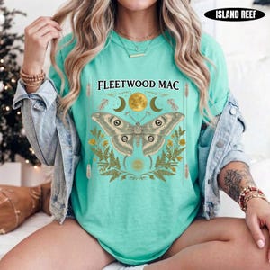 Może przedstawiać: Miętowy t-shirt z grafiką przedstawiającą dużą ćmę nad napisem FLEETWOOD MAC. Wz&oacute;r zawiera złoty księżyc, p&oacute;łksiężyce i liście paproci. Koszulka jest założona pod jasnoniebieską kurtkę dżinsową.
