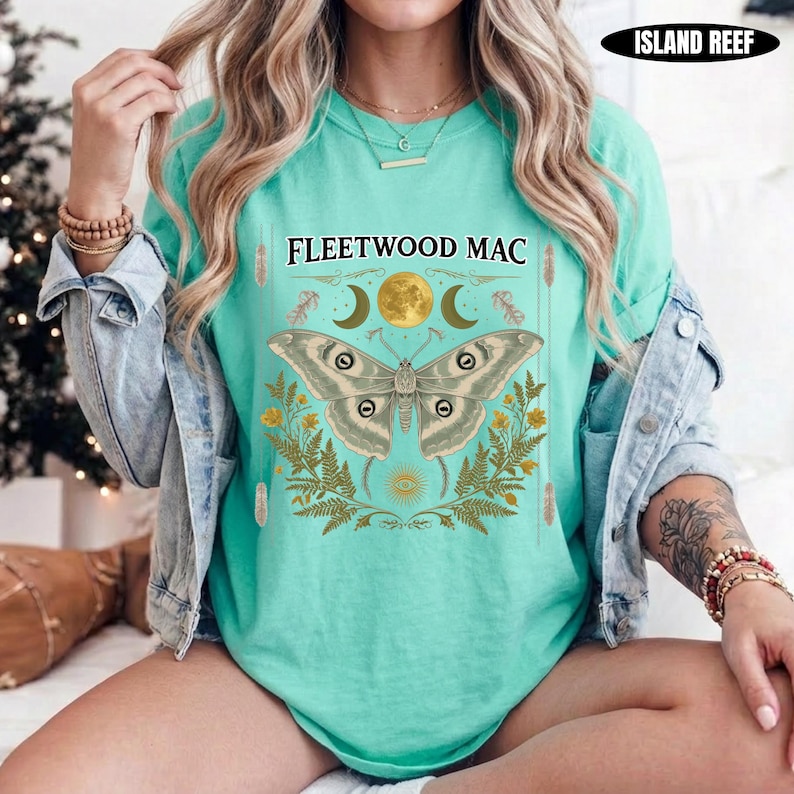 Może przedstawiać: Miętowy t-shirt z grafiką przedstawiającą dużą ćmę nad napisem FLEETWOOD MAC. Wz&oacute;r zawiera złoty księżyc, p&oacute;łksiężyce i liście paproci. Koszulka jest założona pod jasnoniebieską kurtkę dżinsową.