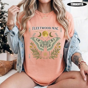 Może przedstawiać: Koszulka w kolorze terakoty z nadrukiem przedstawiającym dużą ćmę umieszczoną nad napisem Fleetwood Mac. Wz&oacute;r zawiera ż&oacute;łty księżyc, p&oacute;łksiężyce i zielone liście paproci. Koszulka jest noszona pod jasnoniebieską kurtką dżinsową.