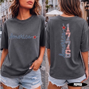 May include: A charcoal grey short-sleeve t-shirt featuring a front graphic with the word America in blue script and a red star. The back graphic displays the word America in blue script above the vertical text 1776 with a red, white, and blue flag pattern.