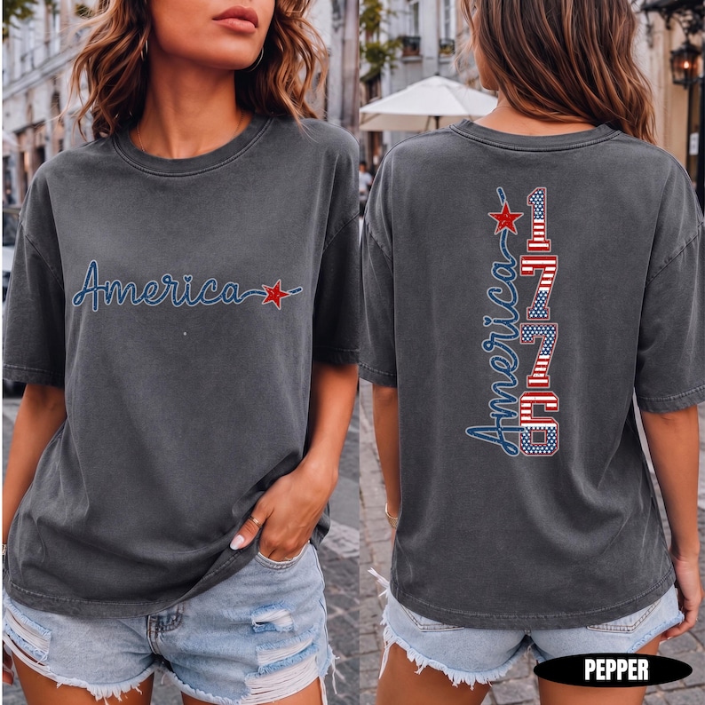 May include: A charcoal grey short-sleeve t-shirt featuring a front graphic with the word America in blue script and a red star. The back graphic displays the word America in blue script above the vertical text 1776 with a red, white, and blue flag pattern.