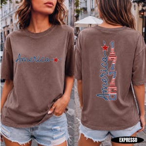 May include: A brown short-sleeved t-shirt featuring the word America in blue script with a red star on the front. The back displays the word America vertically next to the number 1776, both filled with a red, white, and blue flag pattern.