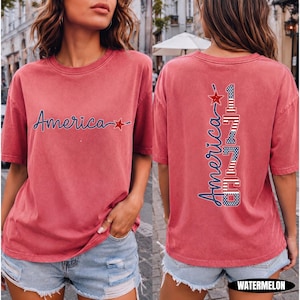 May include: A watermelon-colored short-sleeve t-shirt featuring a front graphic with the word America in blue script and a red star. The back graphic displays the word America vertically next to the number 1776 in red, white, and blue flag patterns.
