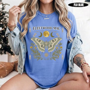 Może przedstawiać: Niebieski t-shirt z napisem FLEETWOOD MAC nad grafiką przedstawiającą dużą ćmę, ż&oacute;łty księżyc i p&oacute;łksiężyce w otoczeniu liści paproci. Koszulka jest noszona pod jasnoniebieską kurtką dżinsową, z widoczną etykietą kolorystyczną FLO BLUE.