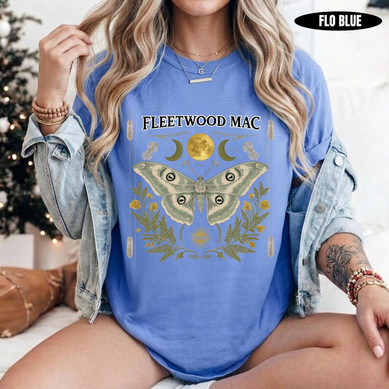 Może przedstawiać: Niebieski t-shirt z napisem FLEETWOOD MAC nad grafiką przedstawiającą dużą ćmę, ż&oacute;łty księżyc i p&oacute;łksiężyce w otoczeniu liści paproci. Koszulka jest noszona pod jasnoniebieską kurtką dżinsową, z widoczną etykietą kolorystyczną FLO BLUE.