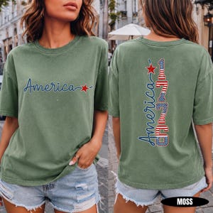 May include: A moss green short-sleeve t-shirt featuring the word America in blue script with a red star on the front. The back displays the word America in blue script vertically alongside the number 1776 in a red, white, and blue flag pattern.