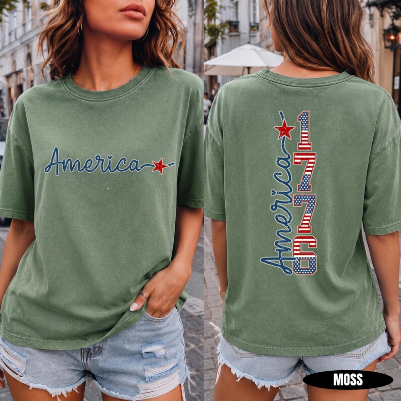 May include: A moss green short-sleeve t-shirt featuring the word America in blue script with a red star on the front. The back displays the word America in blue script vertically alongside the number 1776 in a red, white, and blue flag pattern.