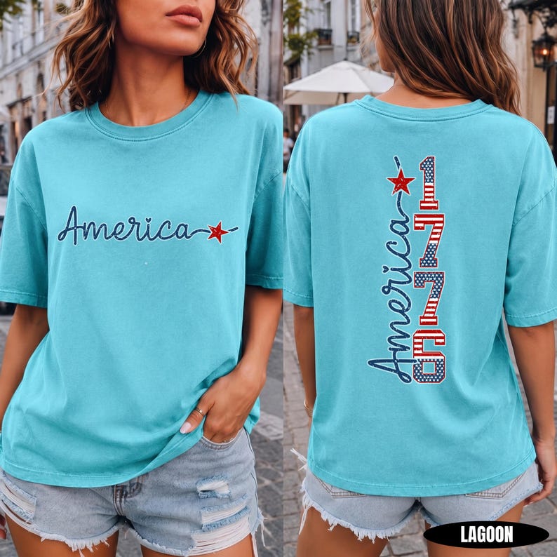 May include: A turquoise short-sleeve t-shirt featuring the word America in dark blue script with a red star on the front. The back displays the word America vertically next to the number 1776, styled with red and white stripes and blue stars.