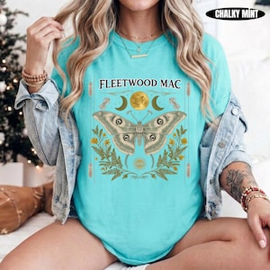 Może przedstawiać: Jasnoturkusowy t-shirt z nadrukiem graficznym przedstawiającym dużą ćmę umieszczoną centralnie nad liśćmi paproci i ikoną słońca. Napis FLEETWOOD MAC jest nadrukowany na czarno nad ćmą, z symbolami p&oacute;łksiężyc&oacute;w po obu stronach.