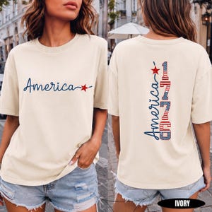 May include: An ivory short-sleeve t-shirt featuring the word America in blue script with a red star on the front. The back displays the word America in blue script above the number 1776, which is filled with a red and white striped pattern.