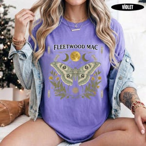 Może przedstawiać: Fioletowy t-shirt z kr&oacute;tkim rękawem z napisem Fleetwood Mac nad grafiką przedstawiającą dużą ćmę, ż&oacute;łty księżyc i zielone liście paproci. Koszulka jest założona pod jasnoniebieską kurtkę dżinsową z metalowymi guzikami i widocznymi szwami.